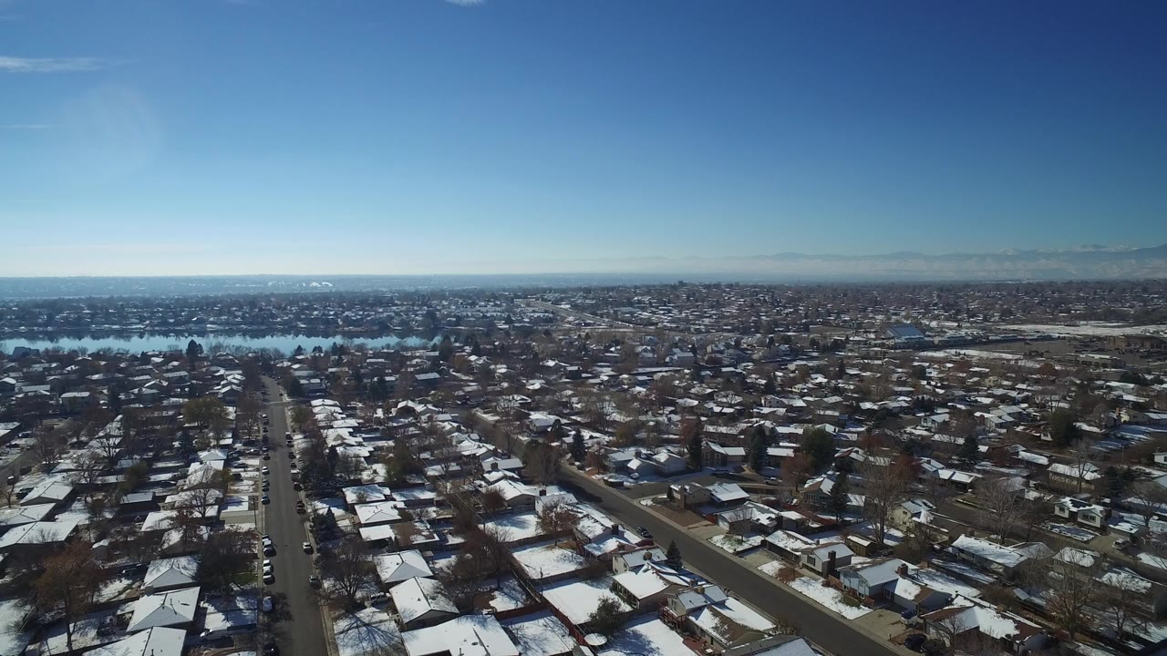 Opening aerial frame over Hidden Lake, Colorado, with snow-dusted houses, a straight residential road, a lake in the midground, distant horizon, and no visible people.