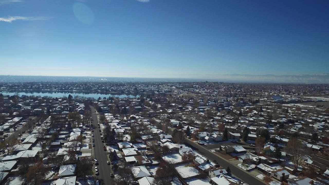 Wide aerial view over snow-covered rooftops and streets in Hidden Lake, Colorado, with a blue lake beyond the neighborhood and no visible people.