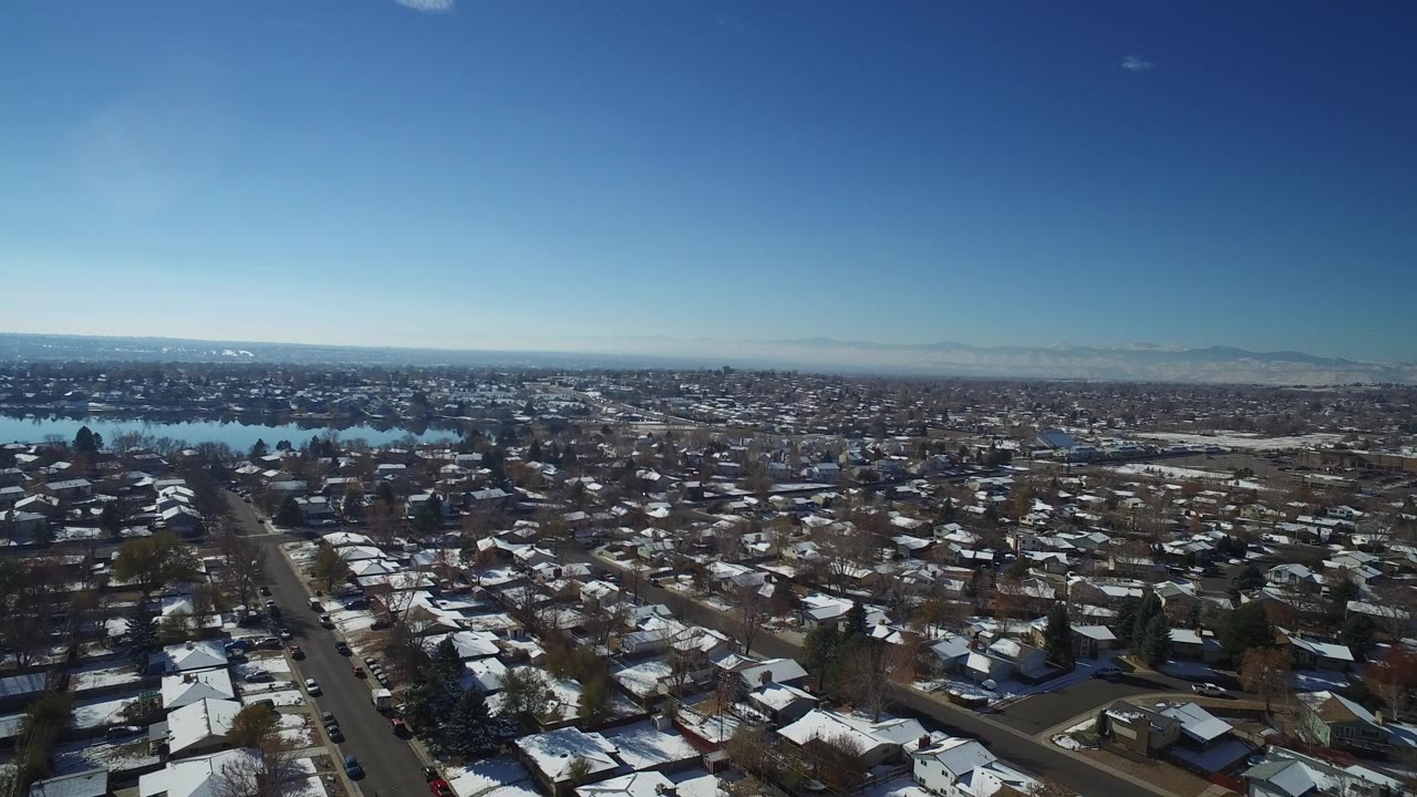 Wide aerial view of snowy residential rooftops beside a lake under clear sky.