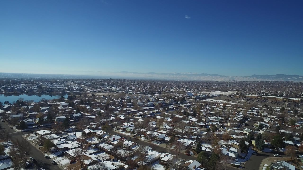 High aerial view over Hidden Lake, Colorado homes with snowy roofs, bare trees, small lakes at left, distant mountains, and no visible people.