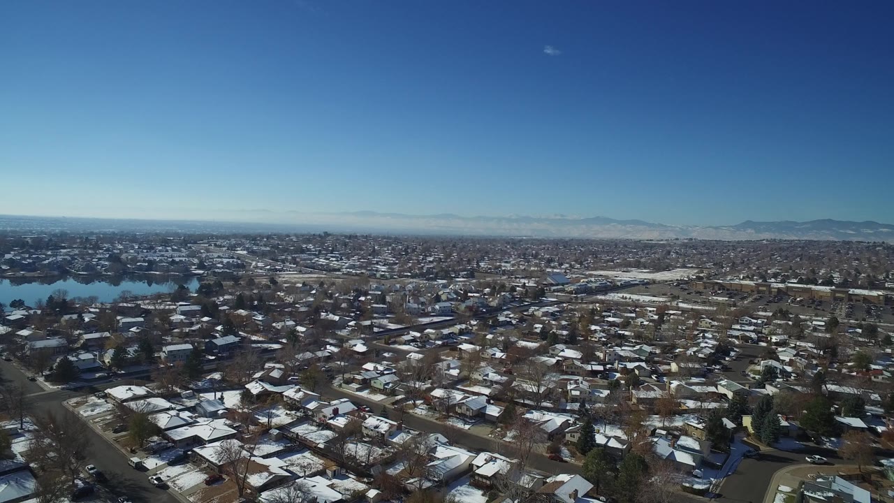 Wide drone-style frame over Hidden Lake, Colorado with residential blocks below, bright roof snow, water in the left midground, mountains on the horizon, and no visible people.
