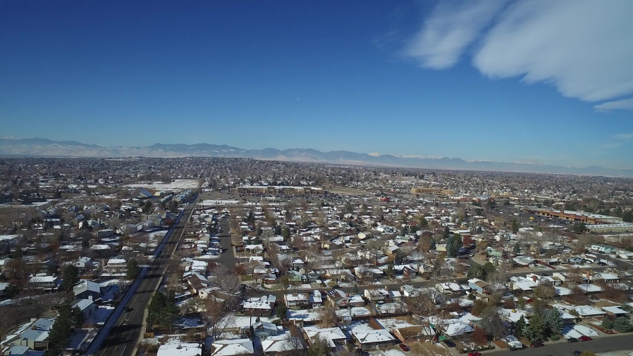 Opening aerial frame over Hidden Lake, Colorado, with snow-dusted rooftops, a straight road at left, distant mountains, blue sky, and no visible people.