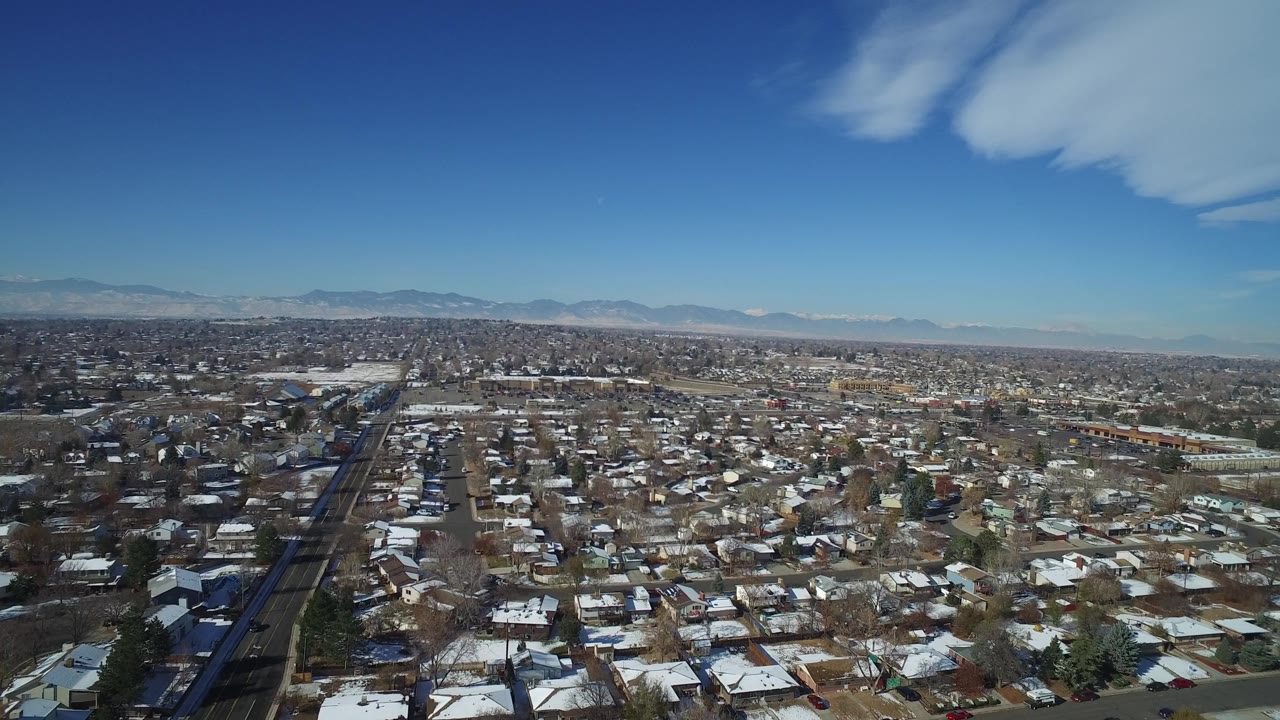Wide Hidden Lake, Colorado aerial with a dark road corridor on the left, dense homes across the frame, pale snow patches, mountains on the horizon, and no visible people.