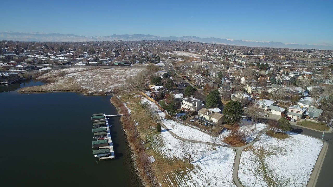 Aerial view over Hidden Lake, Colorado, with dark water at left, snow-covered shoreline lawns, residential homes, docks, distant mountains, and no visible people.