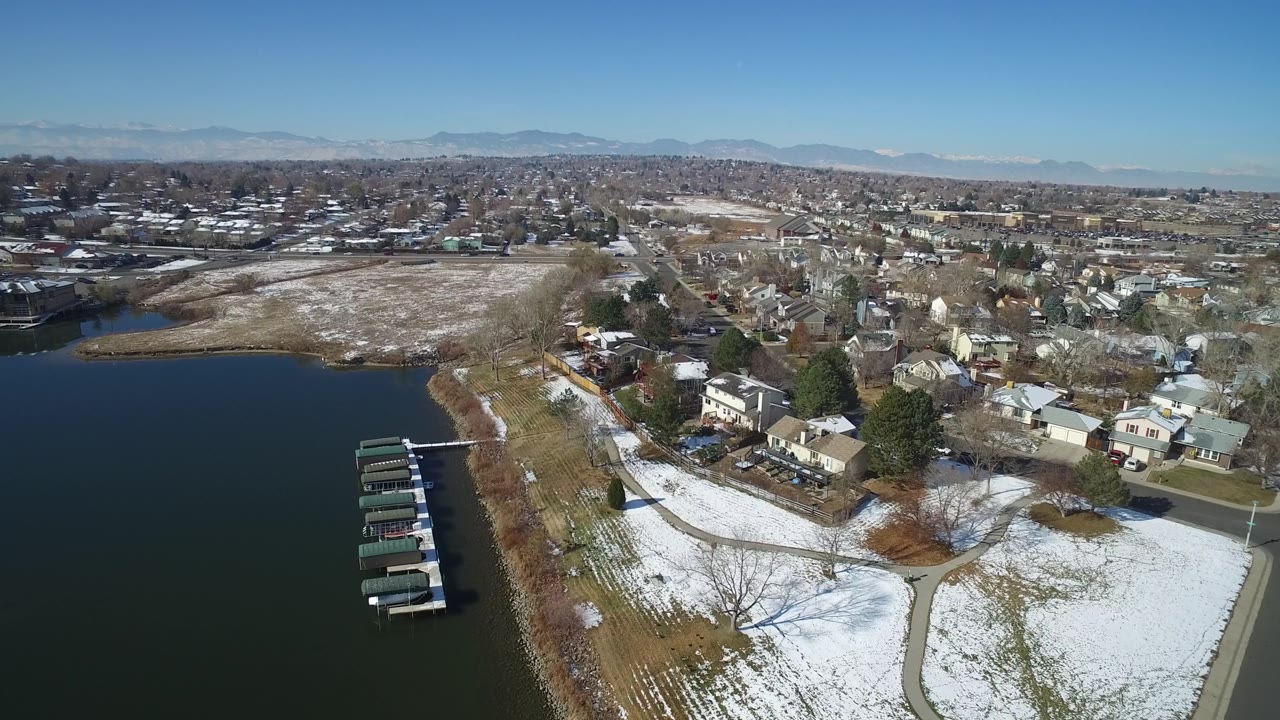 High aerial frame over Hidden Lake, Colorado, with several covered docks near the left shoreline, snowy residential yards to the right, mountains on the horizon, and no visible people.