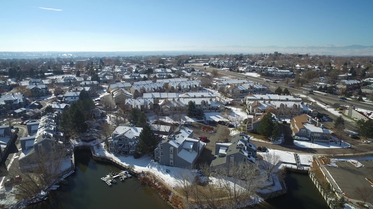 Drone-height view of homes, docks, snow patches, roads, and distant mountains.