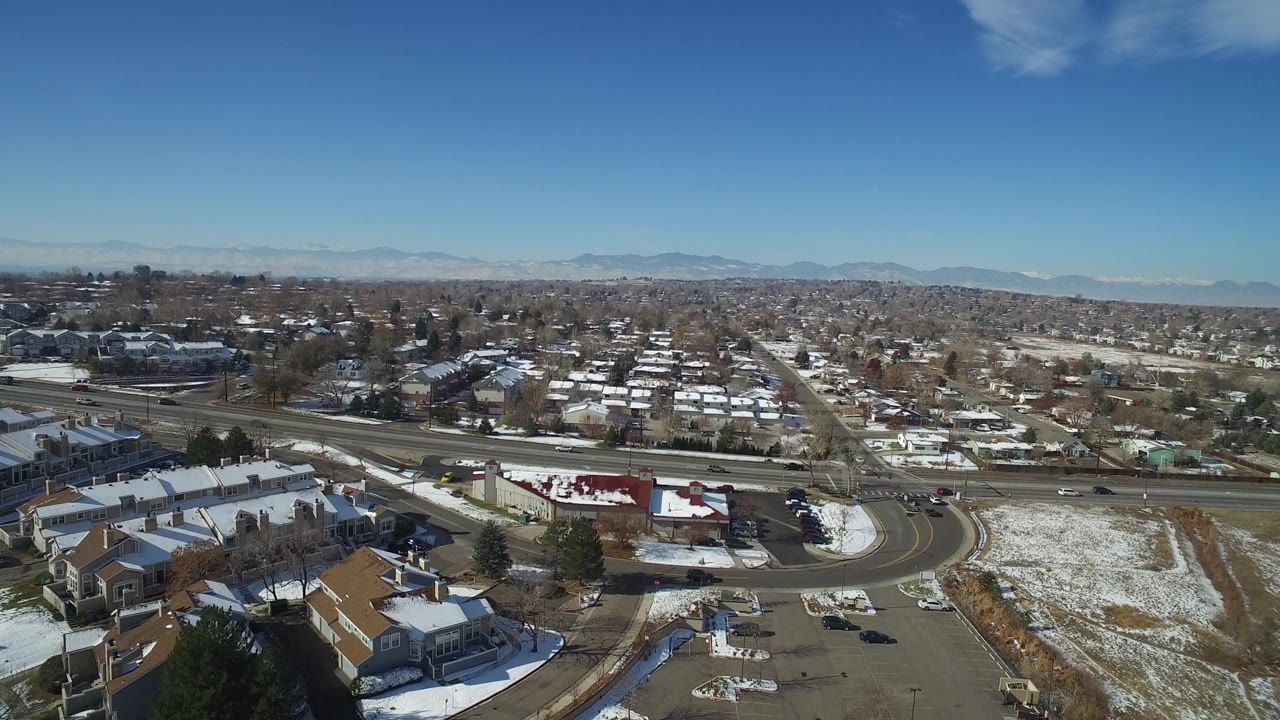 Hidden Lake, Colorado aerial frame showing a red-roofed commercial-looking building, nearby housing blocks, road lanes, snow patches, distant mountains, and no visible people.