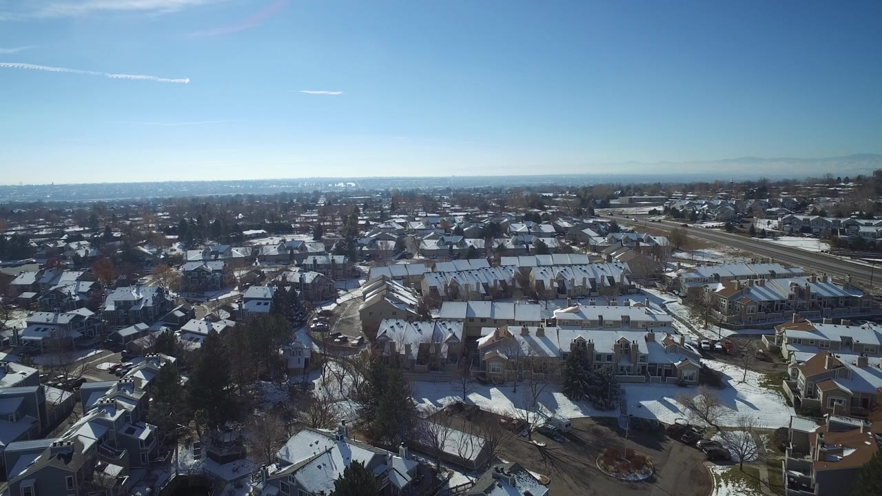 Aerial view over snowy residential rooftops and streets under a blue sky.