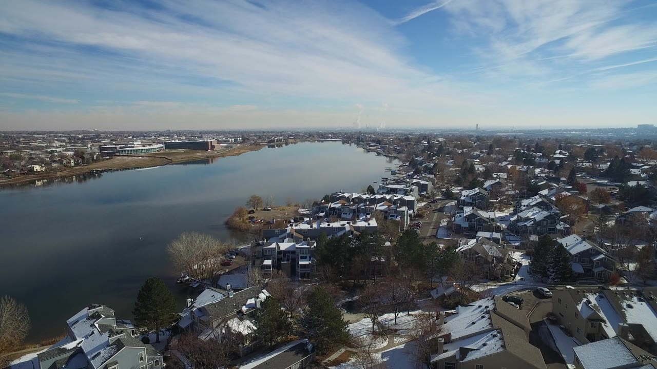 Opening aerial frame over Hidden Lake, Colorado, showing open water, snow-dusted residential roofs, bare trees, pines, distant low buildings, and no visible people.