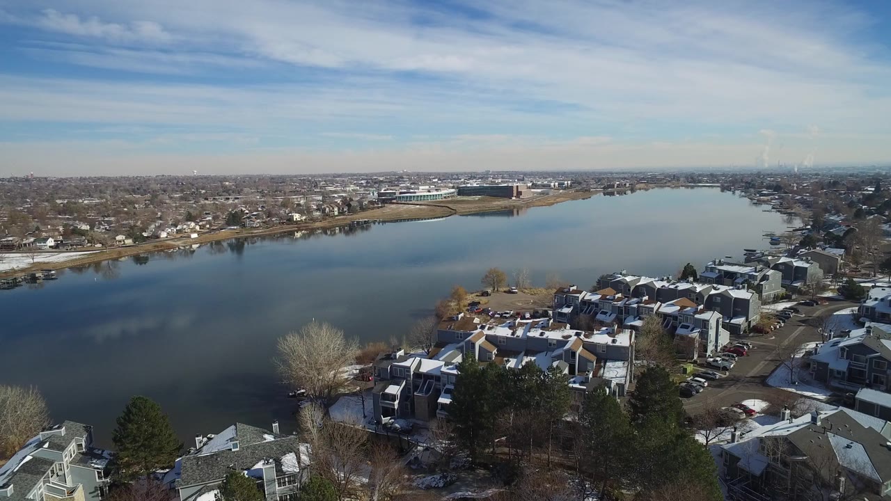 Opening aerial frame over Hidden Lake, Colorado, with snow-dusted townhomes in the foreground, calm water across the center, distant houses on the far shore, and no clearly visible people.