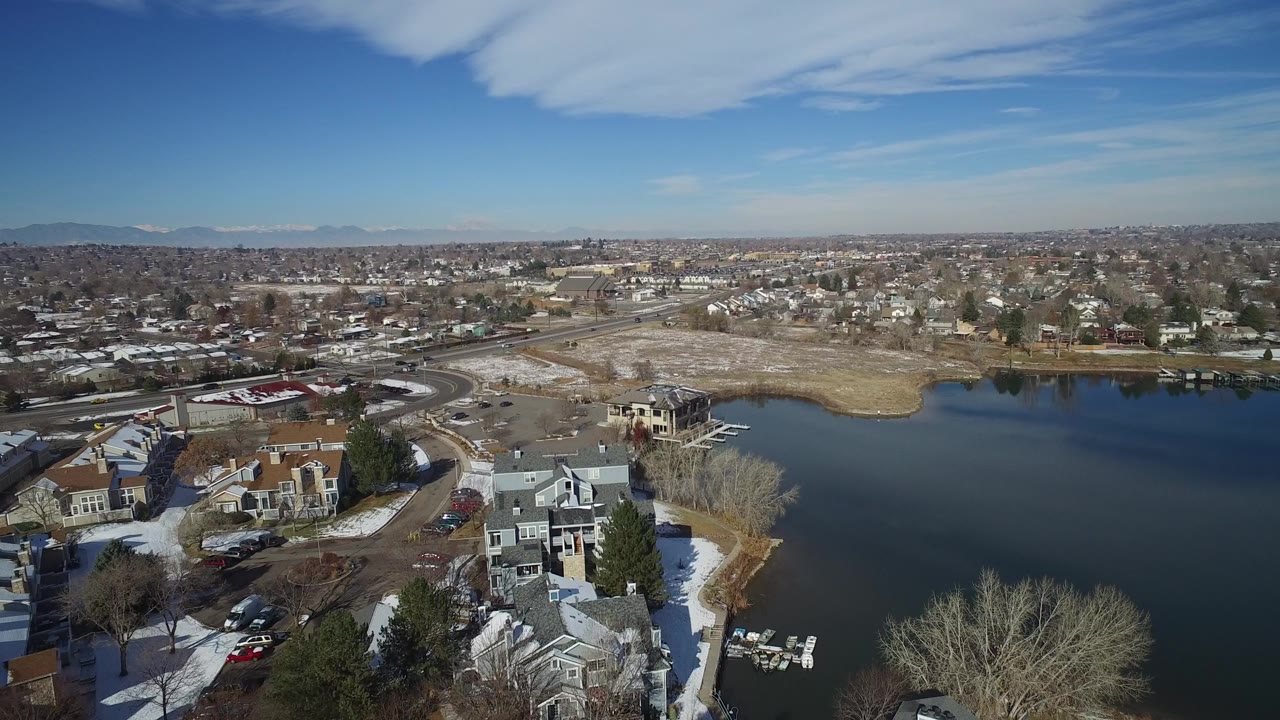Aerial frame over Hidden Lake, Colorado showing dark water, snow-dusted homes, bare trees, roads, distant mountains, blue sky, and no visible people.