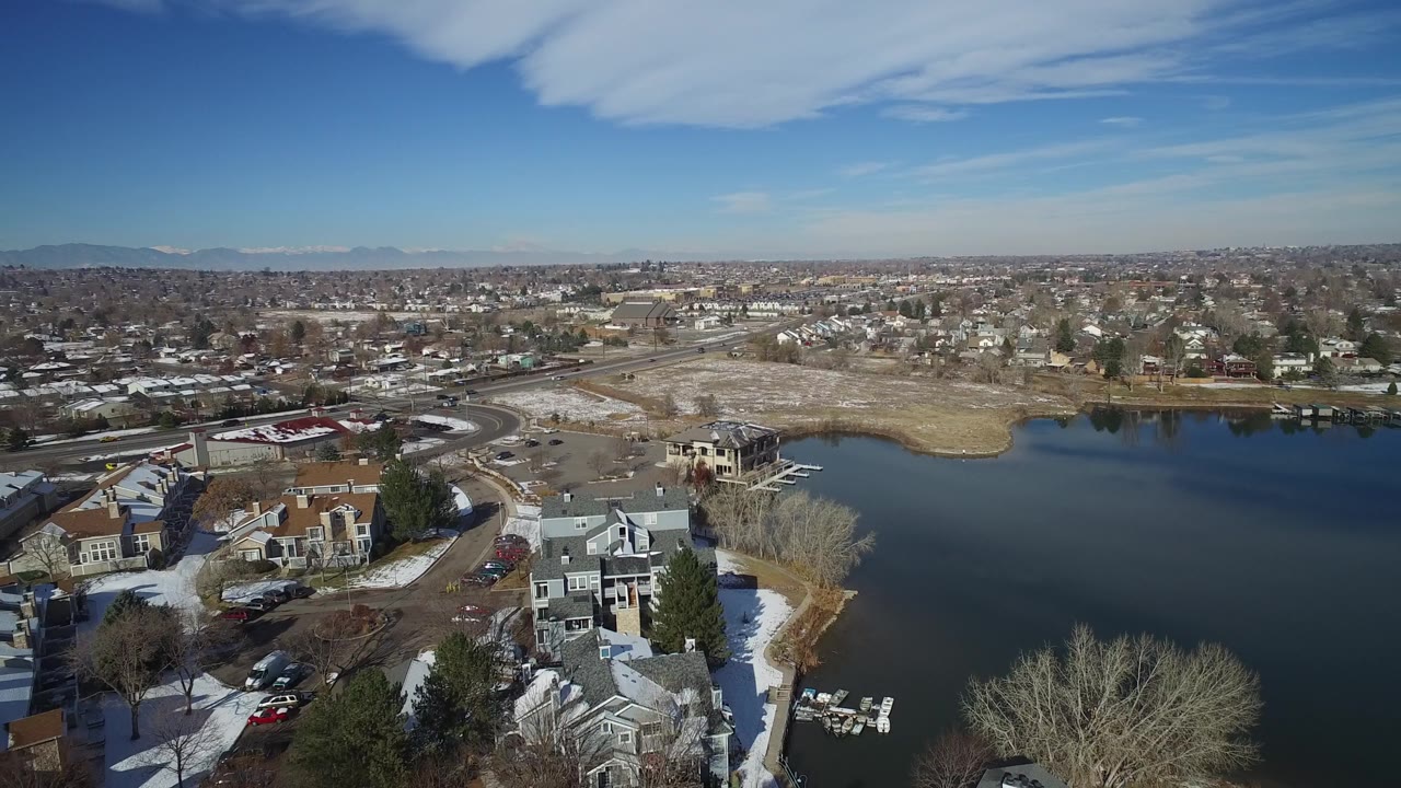 High aerial view in Hidden Lake, Colorado with lakeside houses at the lower edge, open water to the right, roads across the shore, distant mountains, and no visible people.