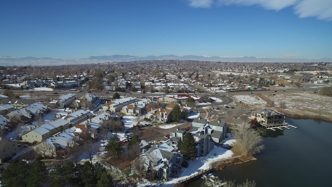 High aerial view over Hidden Lake, Colorado homes beside dark water, with snow on rooftops and lawns, bare trees, distant mountains, and no visible people.