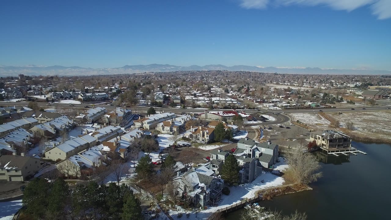 Wide aerial over townhomes, lake water, and winter mountains.