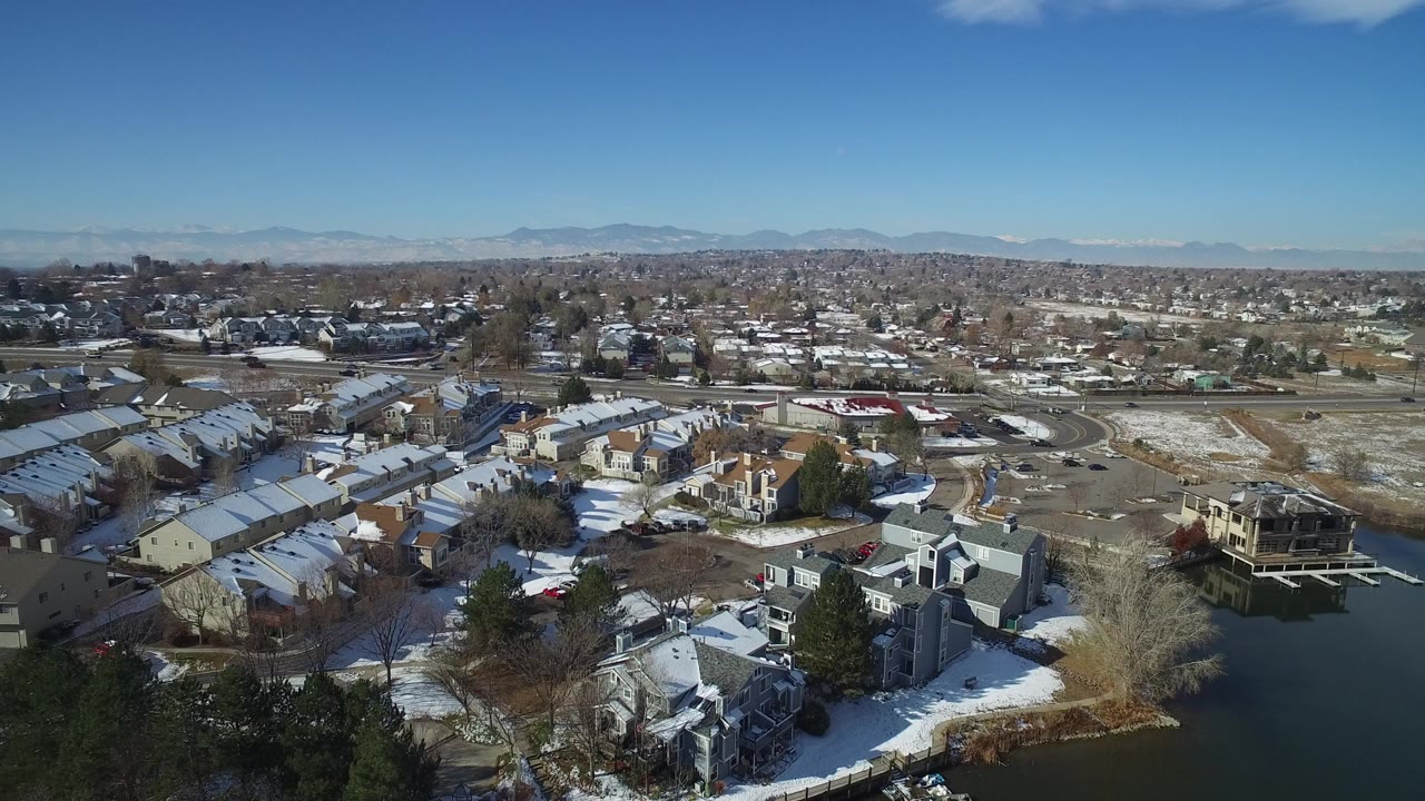 High aerial view over snowy townhomes and suburban streets under a blue sky.