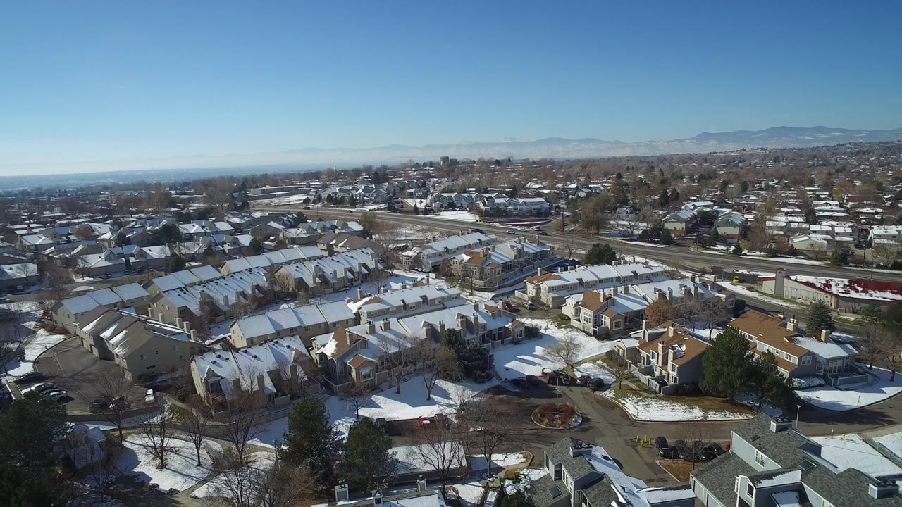 Aerial frame over Hidden Lake, Colorado homes with snow on rooftops and lawns, bare trees, parked cars, distant mountains, and no visible people.