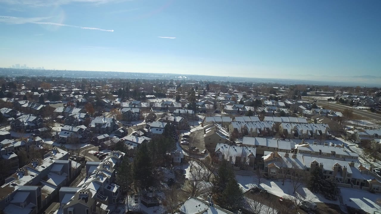 Opening aerial frame over Hidden Lake, Colorado, with snow-dusted rooftops, curving streets, bare trees, distant city buildings, and no visible people.