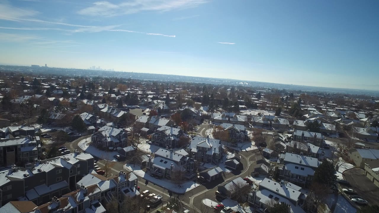 Aerial view over snowy suburban rooftops and curved streets under blue sky.