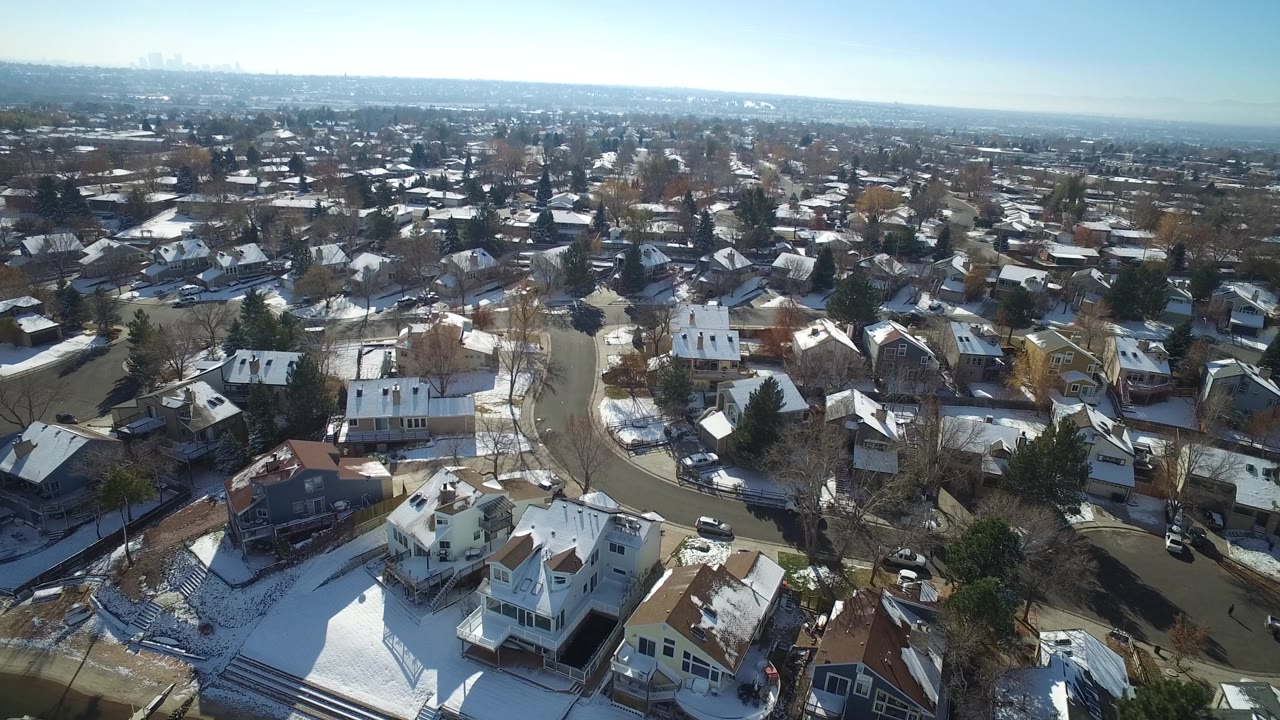 Second aerial frame in Hidden Lake, Colorado with a white house near the lower center, patchy snow on lawns, bare trees, parked vehicles, and no visible people.