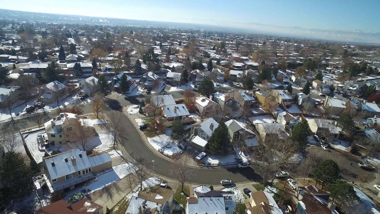 Drone-height view of Hidden Lake, Colorado houses clustered around a curved road, with patchy snow, leafless trees, parked vehicles, and no clearly visible people.