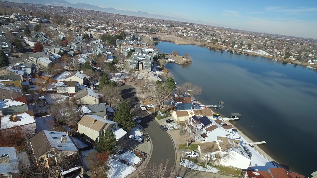 Aerial frame over Hidden Lake, Colorado, with foreground houses and docks, a wide blue lake in midframe, distant residential blocks, and no visible people.