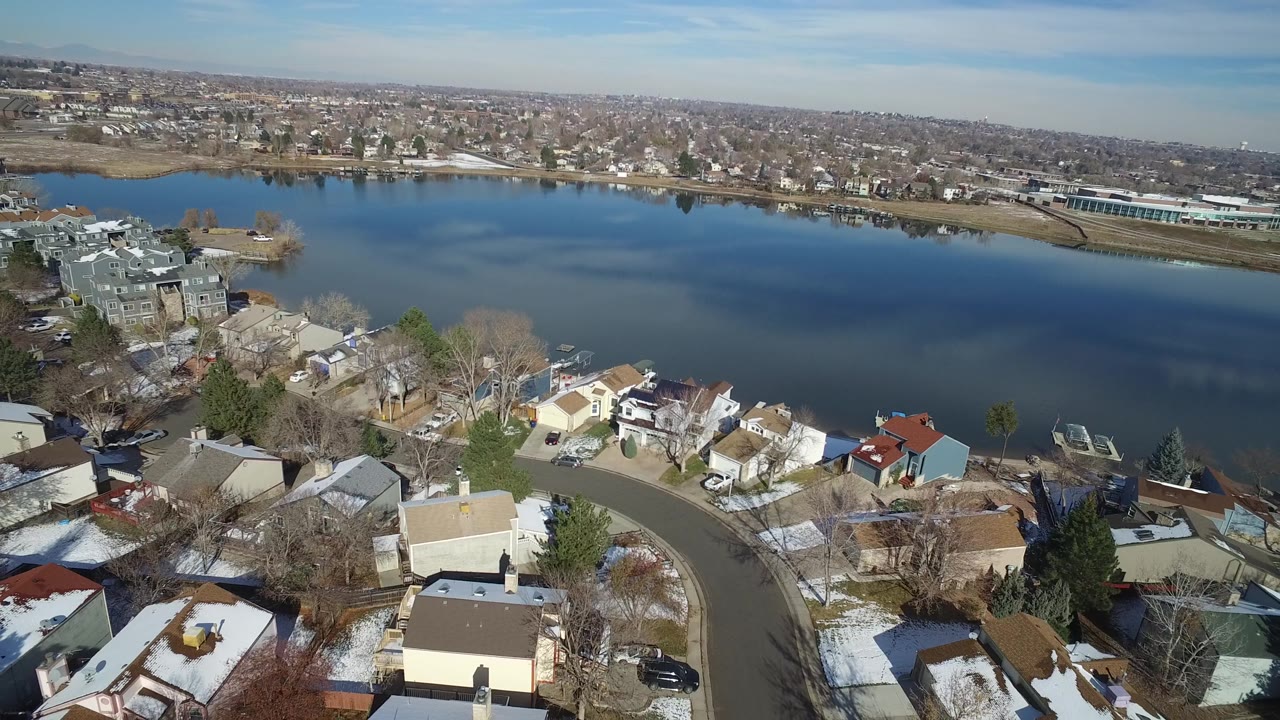 Hidden Lake, Colorado appears from above with foreground rooftops, a bending residential road, bare trees, and the lake stretching across the center of the frame.