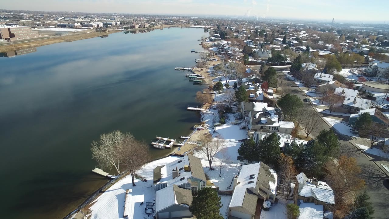 A high aerial view over Hidden Lake, Colorado shows dark water at left, snow-covered homes and docks along the right shoreline, distant buildings on the horizon, and no visible people.