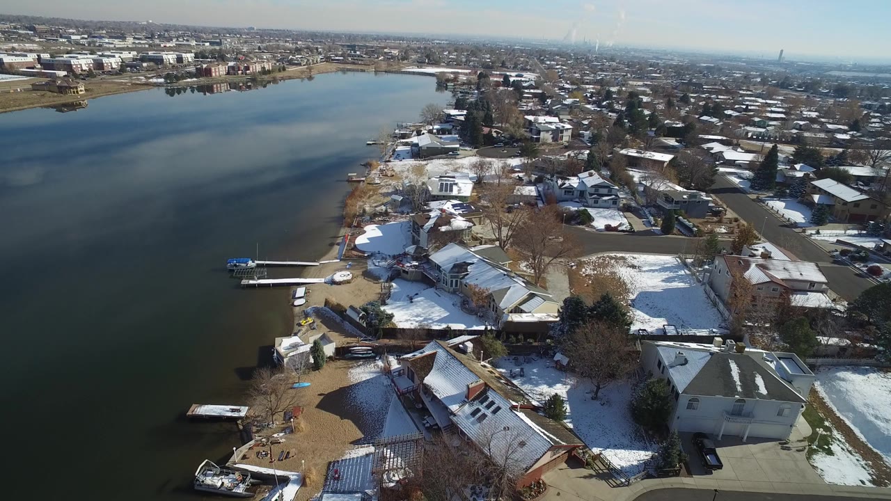High aerial frame over Hidden Lake, Colorado, with dark water on the left, shoreline homes and docks below, winter snow patches on rooftops and yards, and no visible people.