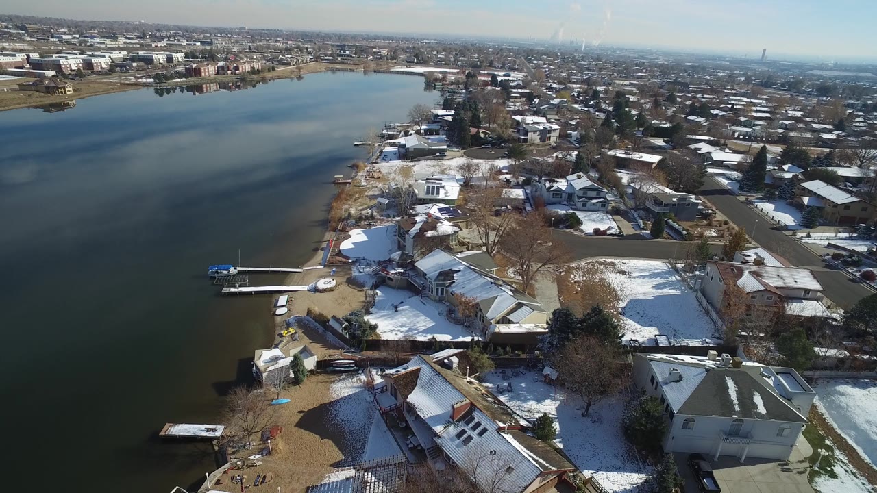 Top view of a residential lakeshore with houses, docks, snow, and distant buildings.