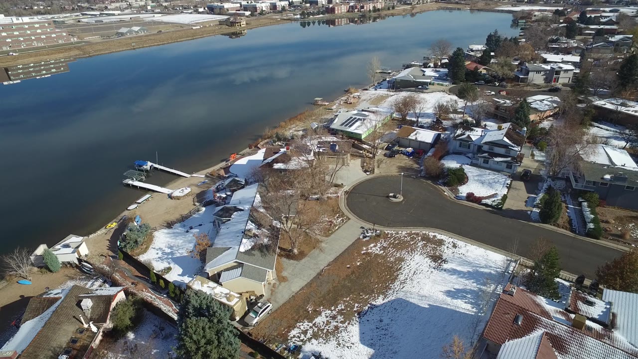 Aerial neighborhood frame with a lake behind homes and snow on roofs and lawns.