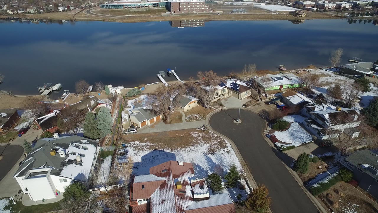 High aerial frame over Hidden Lake, Colorado showing private homes, a curved cul-de-sac, patchy snow, docks, small boats, and a dark waterway, with no visible people.
