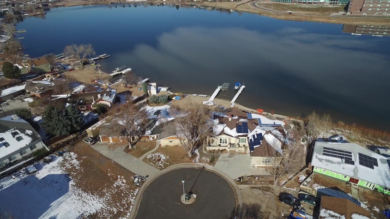 Opening aerial frame over Hidden Lake, Colorado, showing lakeside houses, docks, a cul-de-sac, patchy snow, and calm reflective water with no visible people.