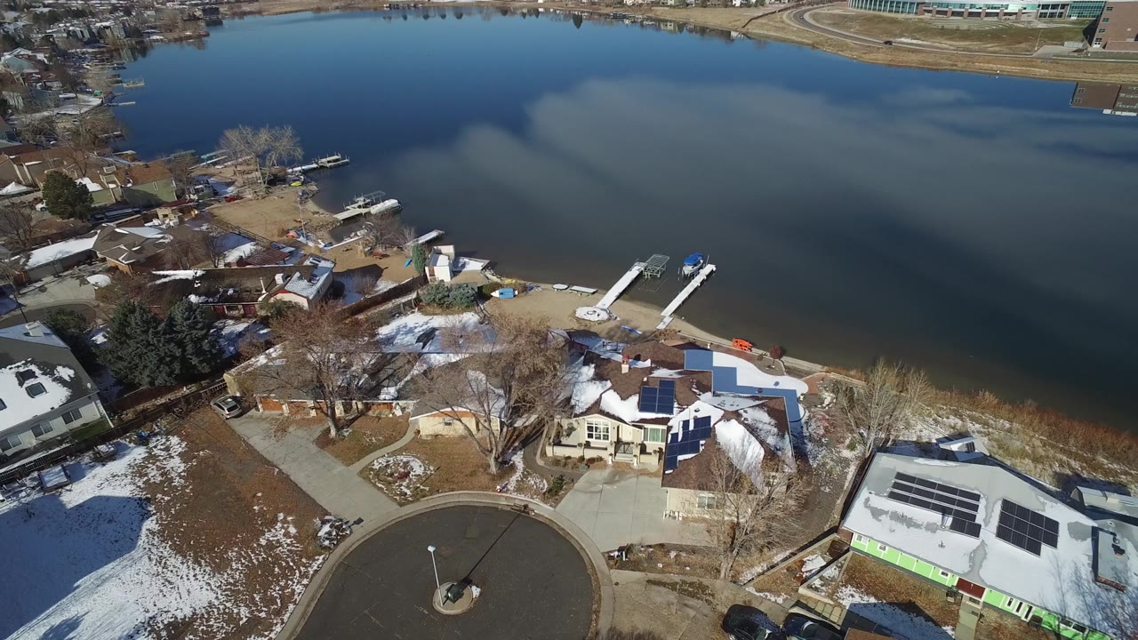 High angle over lakeside houses, snowy lawns, docks, and calm dark water.