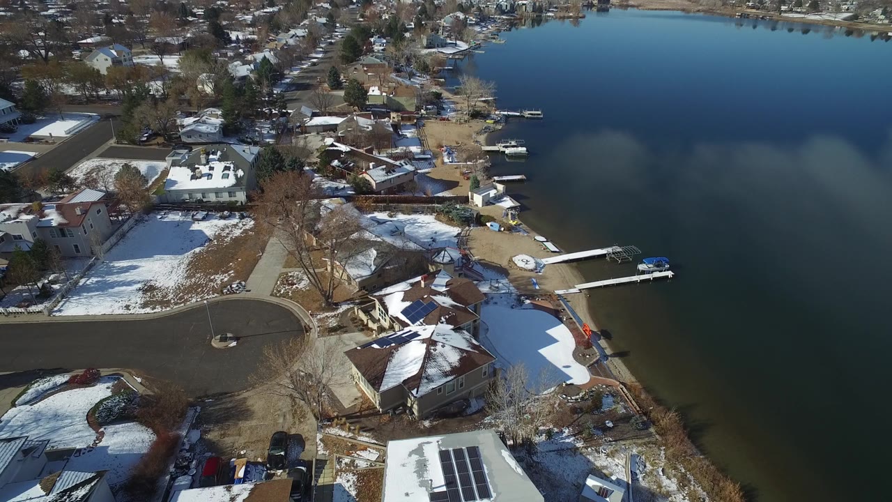 Oblique drone view in Hidden Lake, Colorado with waterfront houses, patchy snow, bare trees, docks, and broad dark water occupying the right side, with no visible people.