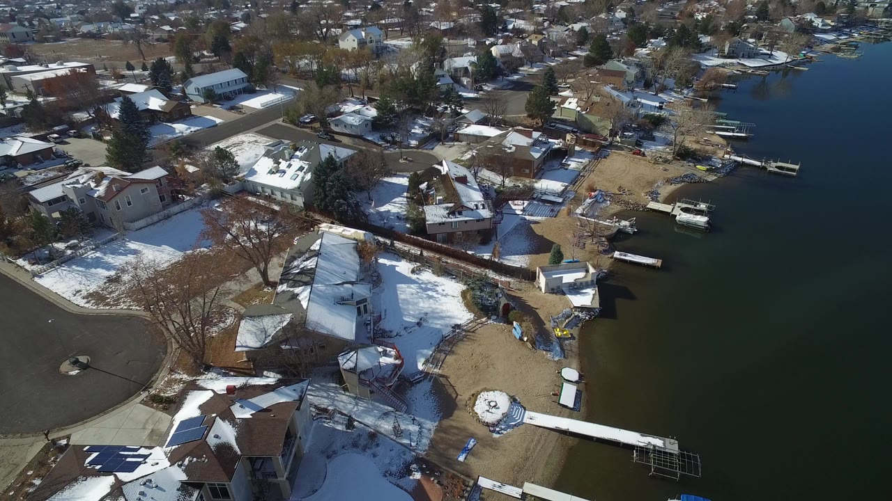 High aerial view over Hidden Lake, Colorado homes with snow on roofs and yards, docks along dark water, leafless trees, and no visible people.