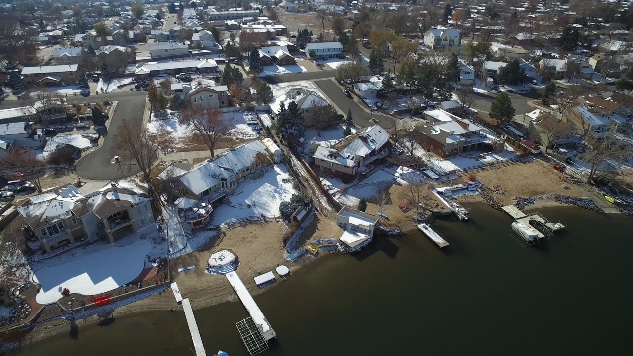 Aerial frame in Hidden Lake, Colorado with lake water at the bottom, several docks, backyards with patchy snow, rooftops, and leafless trees with no visible people.