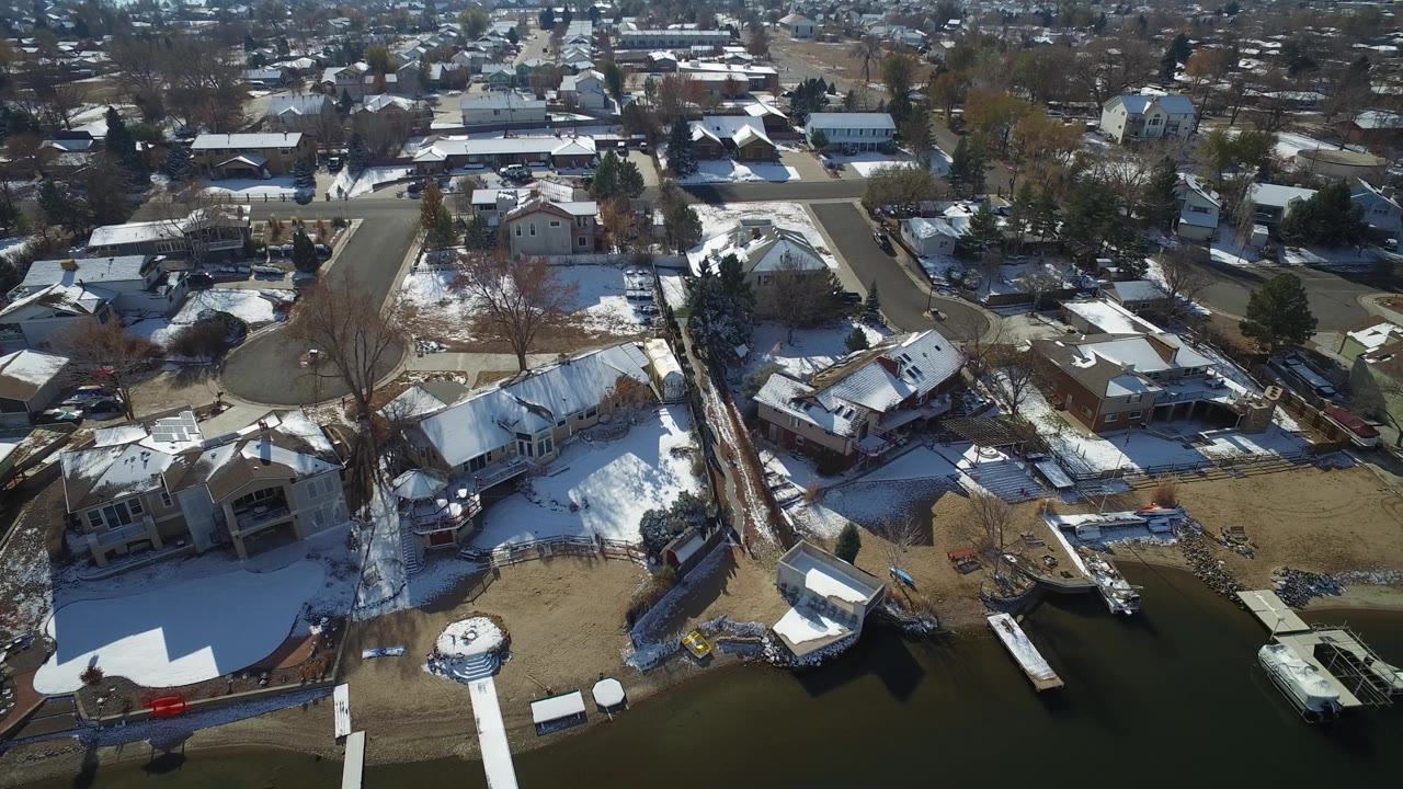 High aerial frame in Hidden Lake, Colorado with a curved residential road, snow-covered roofs, leafless trees, backyard fencing, and lake water along the lower edge, with no visible people.