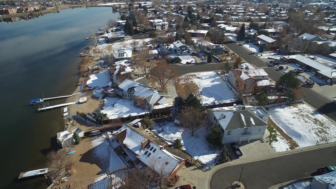 High aerial view over Hidden Lake, Colorado, showing lake water beside detached homes, small docks, paved streets, leafless trees, and patchy snow with no visible people.