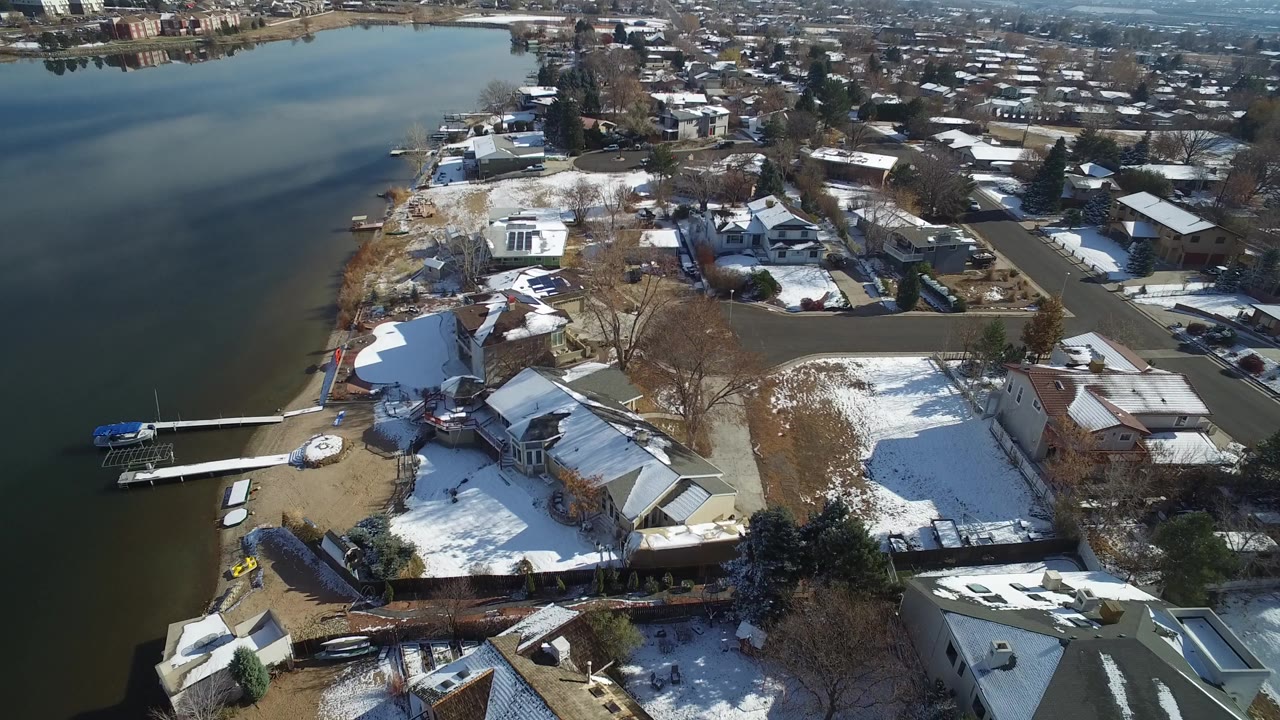 High aerial view over Hidden Lake, Colorado, with a dark lake on the left, waterfront homes, docks, snow-covered lawns, and no visible people.