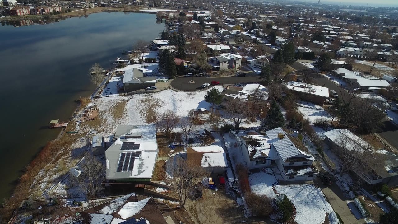 High aerial view over Hidden Lake, Colorado, showing a snow-dusted residential shoreline, rooftop solar panels, leafless trees, pine trees, and dark lake water with no visible people.