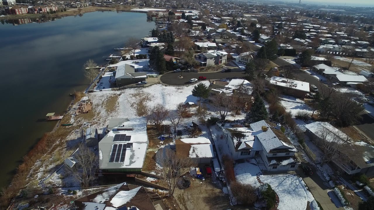 Aerial frame over Hidden Lake, Colorado, with the lake across the upper left, a snowy open yard, a cul-de-sac, private homes, and solar panels on a low roof with no visible people.
