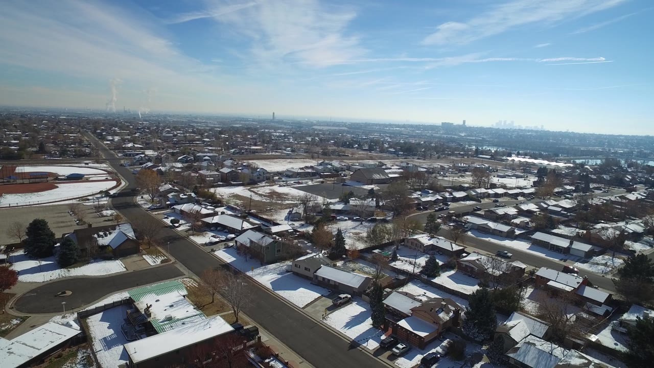 High drone-style view of Hidden Lake, Colorado residential blocks with white snow patches, a broad road in the foreground, distant urban buildings, and no visible people.