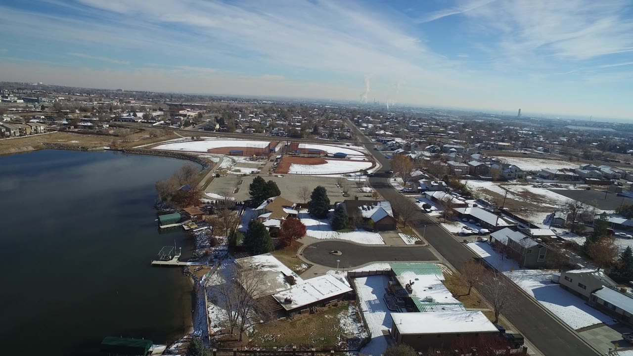 High aerial view in Hidden Lake, Colorado, with the lake at left, snowy lawns and rooftops below, a straight road through the right side, and a wide suburban horizon.