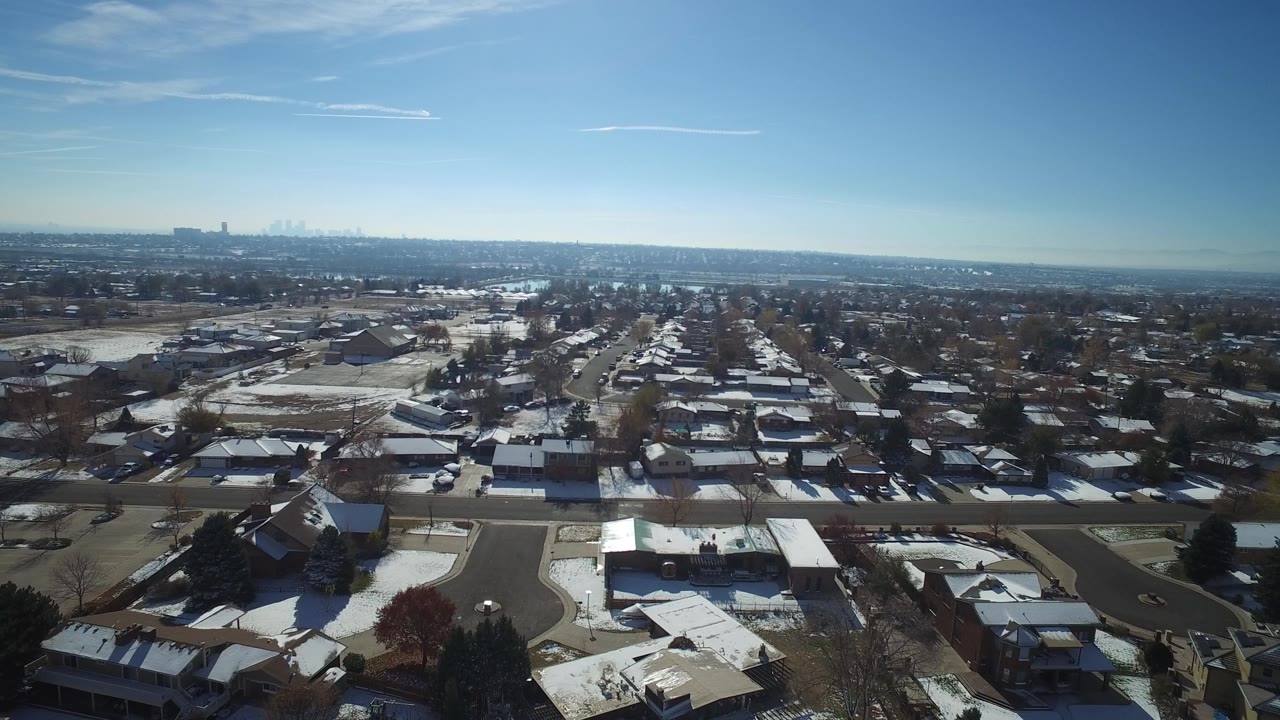 Aerial frame in Hidden Lake, Colorado, looking across rows of single-family homes, bare trees, light snow cover, and a hazy distant urban skyline with no visible people.