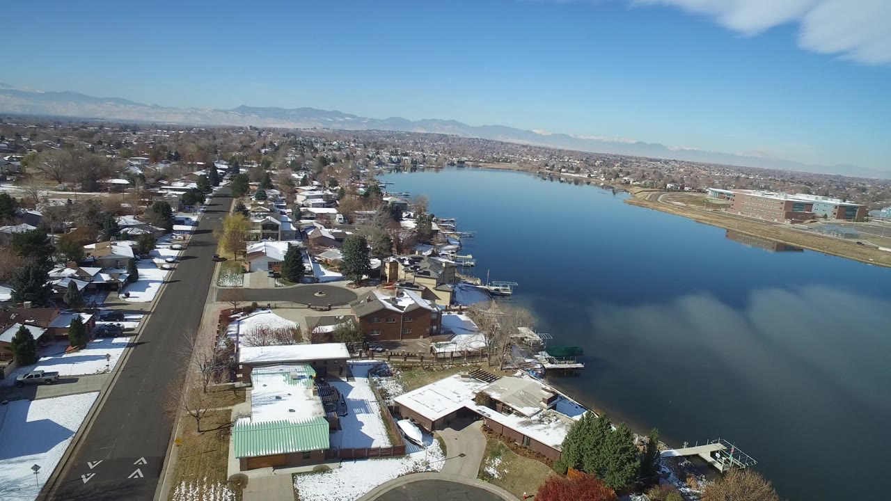Aerial view of lakeside homes, snowy roofs, and a paved street beside calm water.
