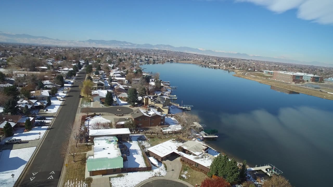 High aerial frame in Hidden Lake, Colorado with a straight residential road on the left, lakeside homes below, small docks on the water, distant urban blocks, mountains, and no visible people.