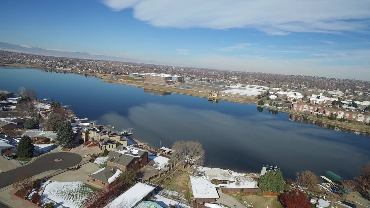 Elevated view over Hidden Lake, Colorado with lakeside homes in the foreground, open water across the middle, distant buildings on the far shore, snow patches, and no visible people.