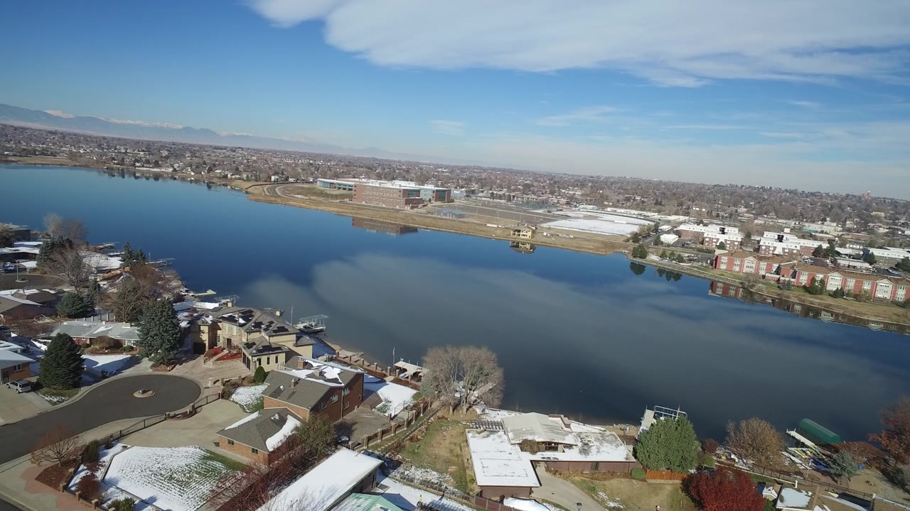 Hidden Lake, Colorado appears from above with a curving residential street and homes below, reflective water in the midground, far-shore buildings, light snow on roofs and lawns, and no visible people.