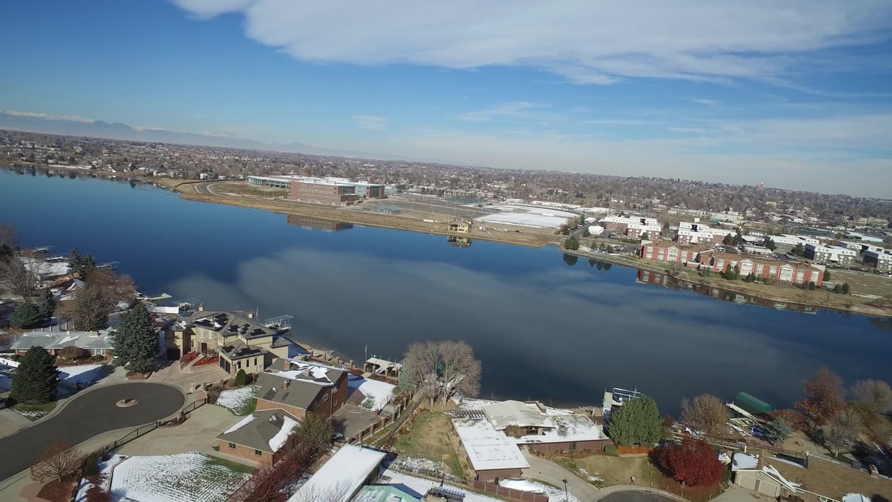 Aerial lakefront neighborhood with snow patches and buildings across the water.