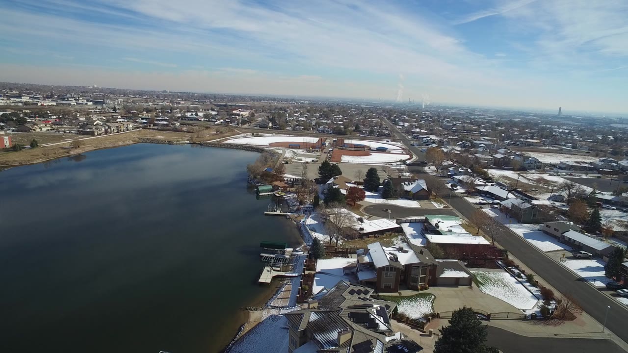 A high aerial view in Hidden Lake, Colorado shows open lake water, snow-dusted lakeside homes, docks, roads, and a distant urban horizon, with no visible people.